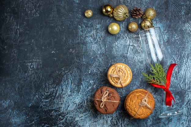 Above view of fallen glass goblet with red ribbon and decoration accessories next to stacked cookies on dark background