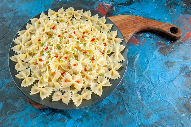 Free photo above view of farfalle pastas with vegetables on a black plate on wooden cutting board on blue background