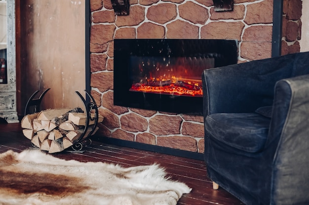 View over fireplace with burning logs, natural fur skin on the floor next to holder with logs in cozy room.