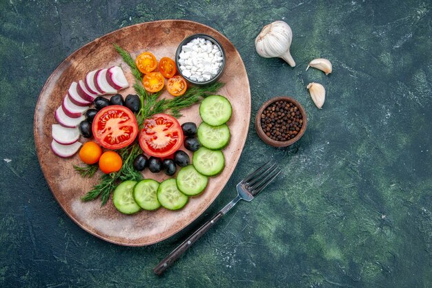 Выше вид свежих нарезанных овощей, оливок в коричневой тарелке и вилки, перца, чеснока на зеленом черном столе смешанных цветов