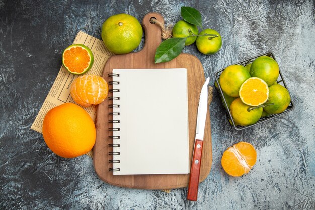 Above view of fresh citrus fruits with leaves notebook on wooden cutting board cut in half forms and knife on newspaper on gray background