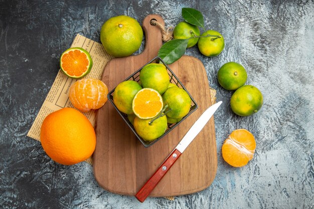 Above view of fresh citrus fruits with leaves on wooden cutting board cut in half forms and knife on newspaper gray table