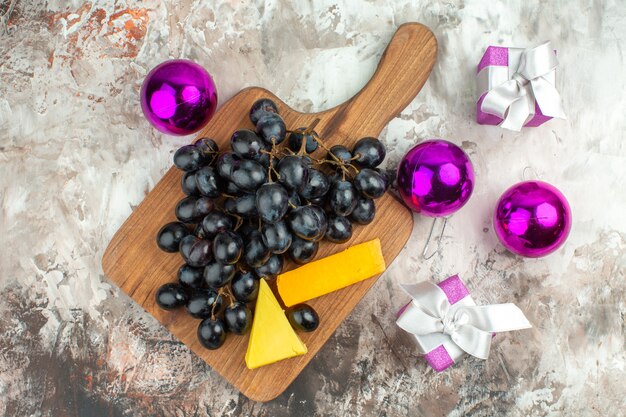 Above view of fresh delicious black grape bunch and cheese on wooden cutting board and gifts decoration accessories on mixed color background