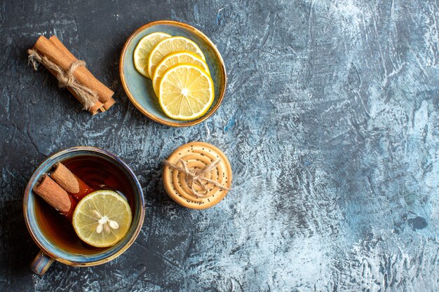 Above view of fresh lemons and a cup of black tea with cinnamon stacked cookies on the right side on dark background
