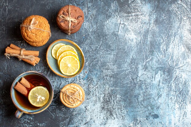 Above view of fresh lemons and a cup of black tea with cinnamon various tacked cookies on the right side on dark background