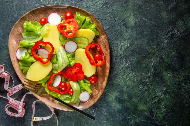 Above view of fresh peeled cut potatoes with red pepper radishes green tomatoes fork in a brown plate and meters on green black mix colors surface