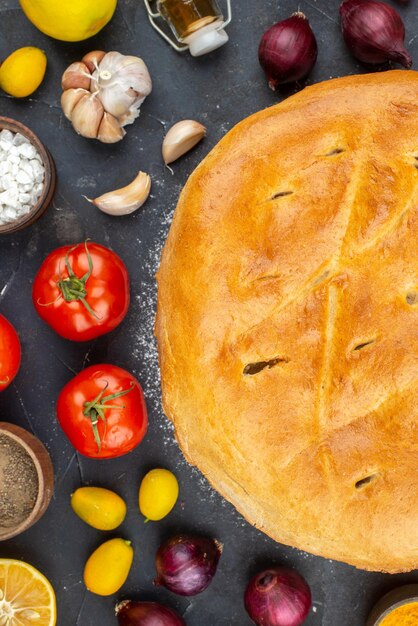 Above view of freshly-baked bread among fresh vegetables fallen oil bottle spices eggs flour on dark color background