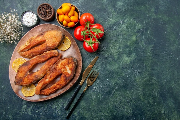 Above view of fried fishes and lemon slices on a brown plate tomatoes kumquats on mix colors table with free space