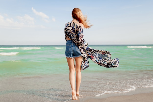 Вид сзади беззаботной грациозной женщины с удивительными рыжими волосами, бегающими по пляжу