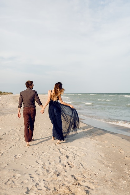 뒤에서 봅니다. 해변을 따라 걷는 사랑에 우아한 커플입니다. 낭만적 인 순간. 하얀 모래와 파도. 열대 휴가. 전체 높이.