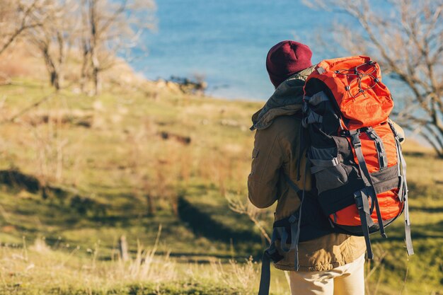 View from back on hipster man traveling with backpack wearing warm jacket and hat, active tourist, using mobile phone, exploring nature in cold season