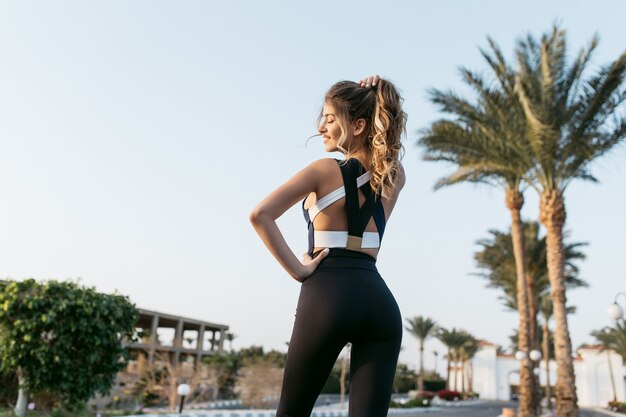 View from back joyful amazing happy woman in sportwear in tropical city. Smiling with closed eyes, sunny morning, palm trees, expressing positivity, true emotions, healthy lifestyle, workout.
