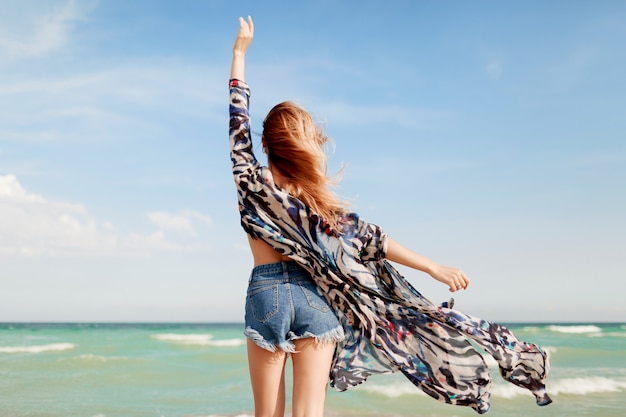 View from back of slim tan red-head girl in stylish tropical outfit posing on amazing beach near ocean