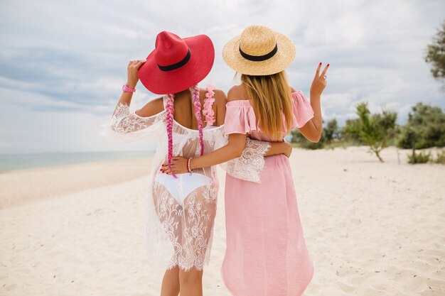 View from back on two beautiful stylish woman at beach on vacation, summer style, fashion trend, wearing straw hats, fashion trend, pink and lace dress, sexy outfit