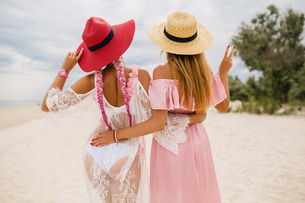Вид сзади на двух красивых стильных женщин на пляже в отпуске, летний стиль, модная тенденция, в соломенных шляпах, модная тенденция, розовое и кружевное платье, сексуальный наряд