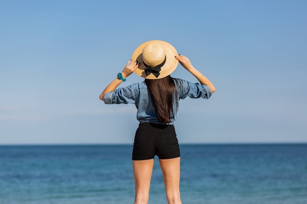 View from back. woman in straw hat, with figure looking on the sea.