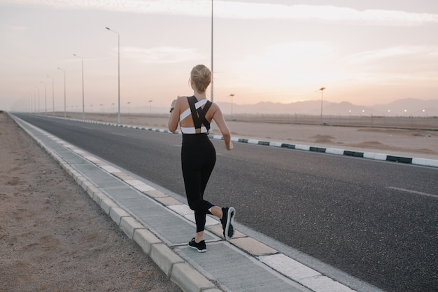 Вид сзади тренировки на дороге привлекательной молодой женщины в спортивной одежде в солнечное утро. Здоровый образ жизни, обучение, сильная спортсменка.