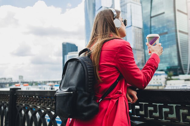 View from back on young hipster woman in pink coat, jeans walking in street with backpack and coffee listening to music on headphones, wearing sunglasses