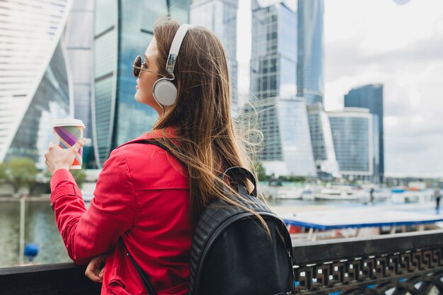 View from back on young hipster woman in pink coat, jeans walking in street with backpack and coffee listening to music on headphones
