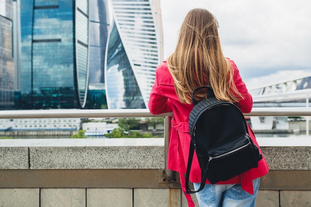 View from back on young hipster woman in pink coat walking on stairs in street with backpack and coffee listening to music on headphones, wearing sunglasses