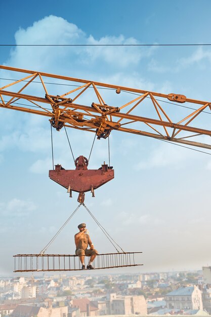 View from distance of biggest crane holding iron construction, where sitting builder and eating. Man resting and looking down. Cityscape on background. Extreme building on high.