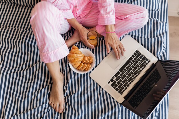 View from above on hands of young woman in pink pajamas sitting on bed with laptop having breakfast