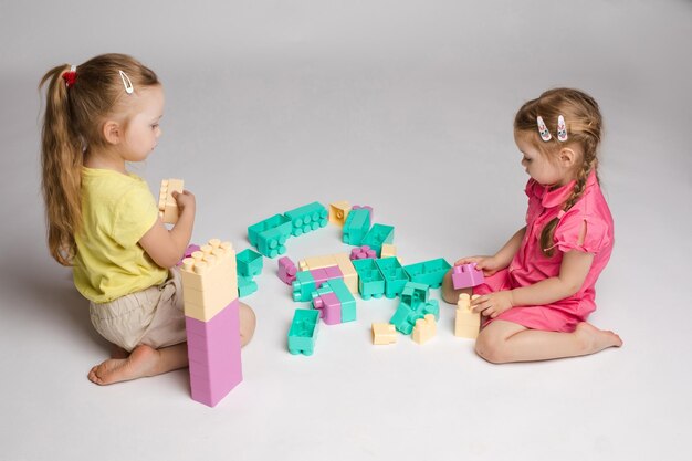 View from side of two cute girls sitting on floor and playing with building blocks in studio Little children enjoying game talking and smiling on isolated background Concept of joy and activity