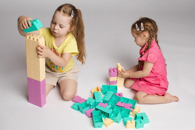 View from side of two cute girls sitting on floor and playing with building blocks in studio Little children enjoying game talking and smiling on isolated background Concept of joy and activity