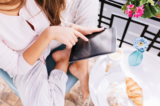 Вид сверху планшет в руках девушки в пижаме, сидящей на балконе, завтракающей.