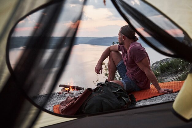 View from tent of traveller with backpack sitting on top of mountain enjoying view coast a river or lake.