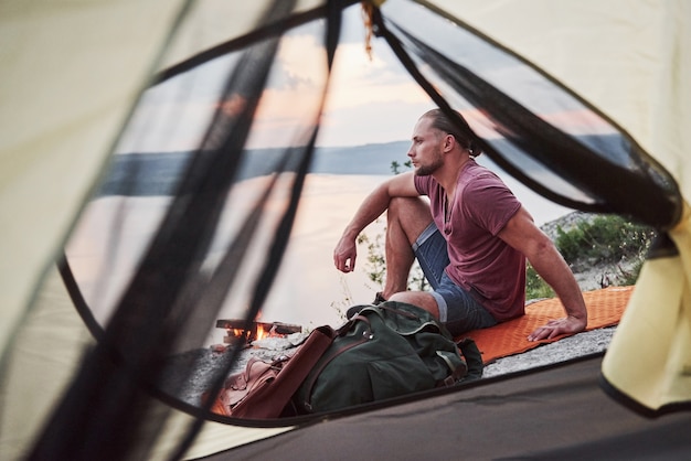 View from tent of traveller with backpack sitting on top of mountain enjoying view coast a river or lake.