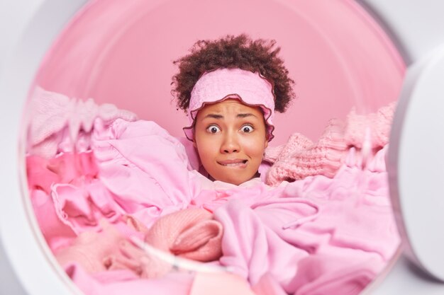 View from washer of nervous housewife with curly hair has anxious expression bites lips drowned in pile of laundry wears headband busy doing housework