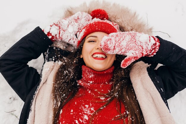 View from above on young pretty candid smiling happy woman in red mittens and knitted hat wearing black coat lying in snow in park, warm clothes, having fun