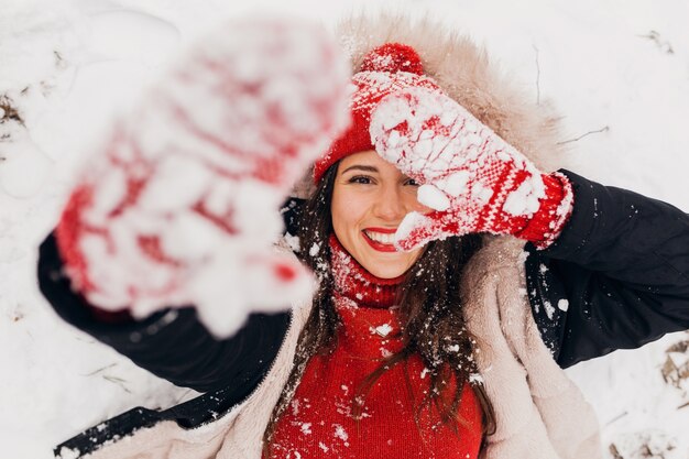 Вид сверху на молодую довольно откровенную улыбающуюся счастливую женщину в красных варежках и вязаной шляпе в черном пальто, лежащую в снегу в парке, теплую одежду, веселую
