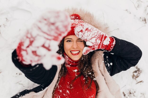 公園で雪の中で横たわっている黒いコートを着て、暖かい服を着て、楽しんで、赤いミトンとニット帽の若いかなり率直な笑顔の幸せな女性を上から見る