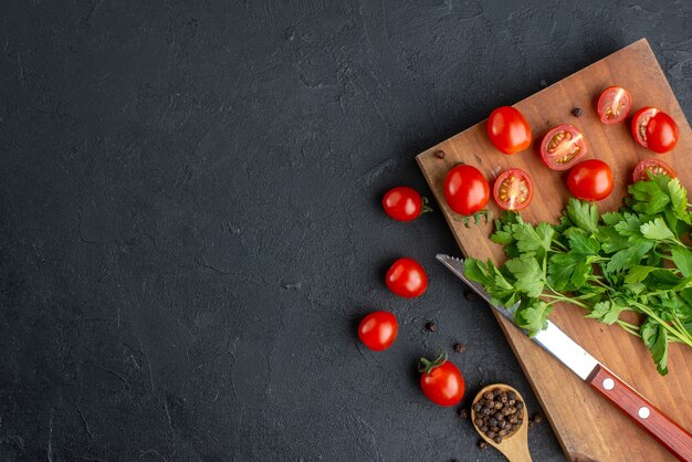 Above view of green bundle fresh whole cut tomatoes on wooden cutting board knife on the left side on black distressed surface