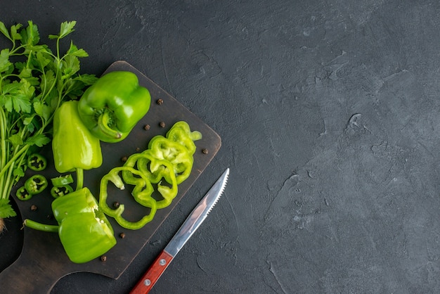 Above view of green bundle fresh whole green peppers on wooden cutting board knife on the right side on black distressed surface