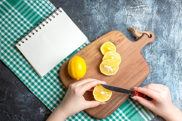 Above view of a hand chopping fresh lemons on a wooden cutting board and notebook on green stripped cloth