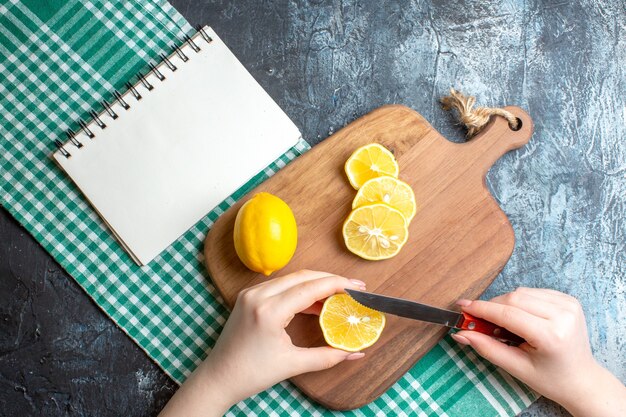 Above view of a hand chopping fresh lemons on a wooden cutting board and notebook on green stripped cloth