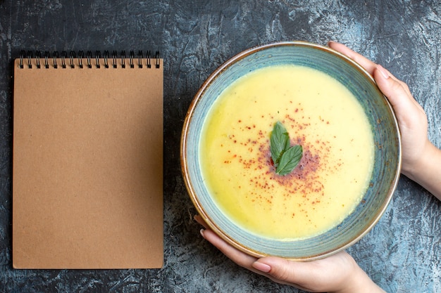 Above view of hand holding a blue pot with tasty soup and spiral notebook on blue background