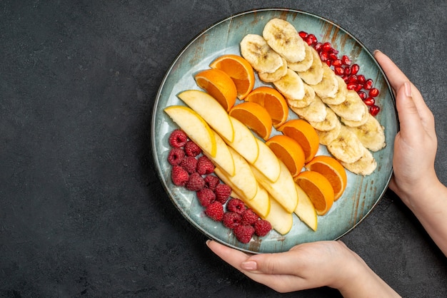 Above view of hand holding collection of chopped fresh fruits on a blue plate on black table