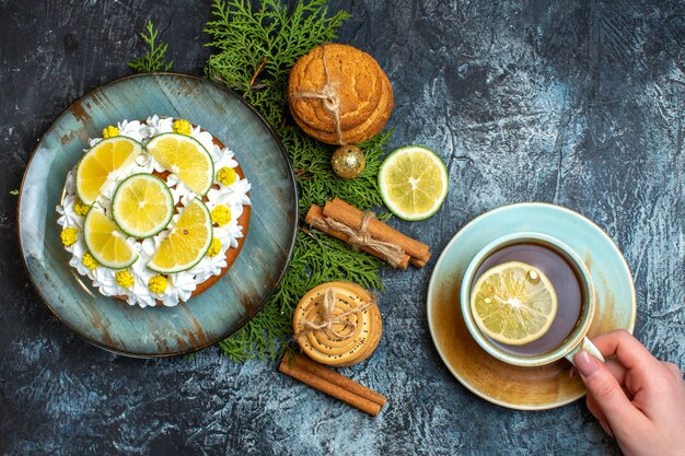 Above view of hand holding a cup of black tea creamy delicious cake and fir branches lemon cinnamon limes on dark background