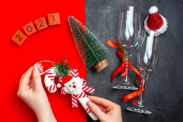 Above view of hand holding decoration accessories glass goblets christmas tree numbers santa claus hat on red and black background
