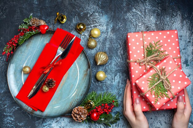 Above view of hand holding gift boxes and cutlery set with red ribbon on a decorative napkin on a blue plate and christmas accessories and christmas sock on dark background