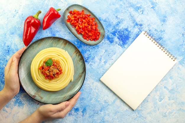 青いテーブルの上のスパイラルノートの横にあるおいしいスパゲッティのみじん切りと赤唐辛子全体と灰色のプレートを持っている手のビューの上