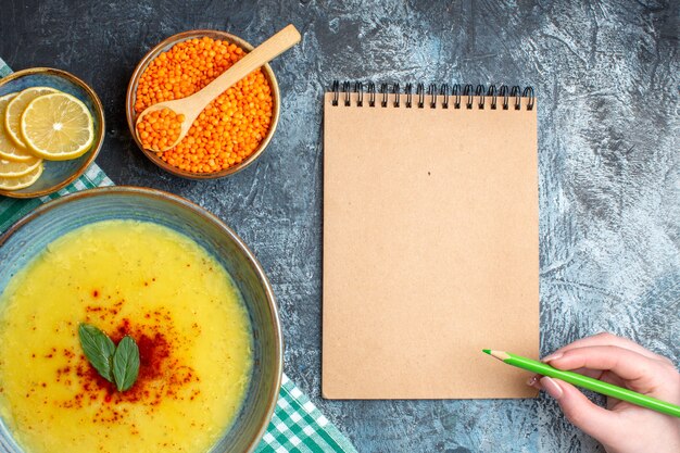 Above view of hand holding pen on spiral notebook tasty soup served with mint and pepper on a green stripped towel