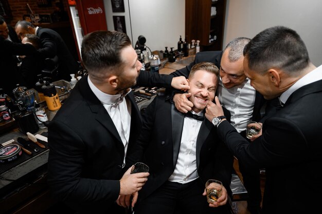Above view of happy and stylish bridegroom in suit sitting in chair and friends surrounded him which holding his cheeks and having fun during wedding preparation in barber shop