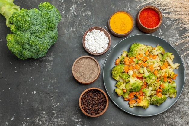 Free photo above view of healthy meal with brocoli and carrots on a black plate and spices on gray table