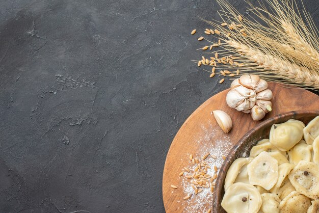 Above view of homemade dumplings in a brown bowl on a wooden cutting board spikes garlics on dark table with free space