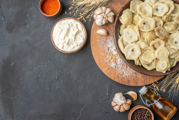 Above view of homemade dumplings in a brown bowl on a wooden cutting board spikes garlics pepper fallen oil bottle on dark table with free space